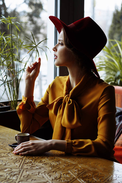 portrait of a girl in a cafe with a cup of coffee and a hat.portrait of sensual young girl wearing floppy hat and blouse with bow. Beautiful brunette woman in cafe holding cup of coffee - 写真・画像