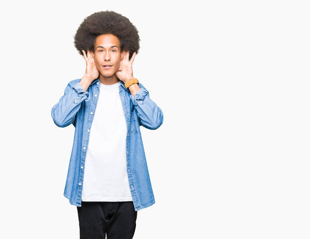 Giovane uomo afro-americano con i capelli afro che cerca di sentire entrambe le mani sul gesto dell'orecchio, curioso di pettegolezzi. Problemi uditivi, sordi
 - Foto, immagini