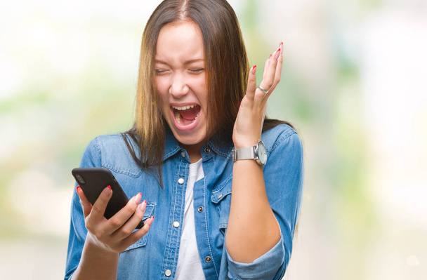 junge schöne kaukasische Frau sendet SMS mit Smartphone über isolierten Hintergrund sehr glücklich und aufgeregt, Siegerausdruck feiert Sieg schreiend mit großem Lächeln und erhobenen Händen - Foto, Bild