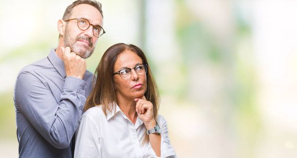 Middle age hispanic couple in love wearing glasses over isolated background with hand on chin thinking about question, pensive expression. Smiling with thoughtful face. Doubt concept. - 写真・画像