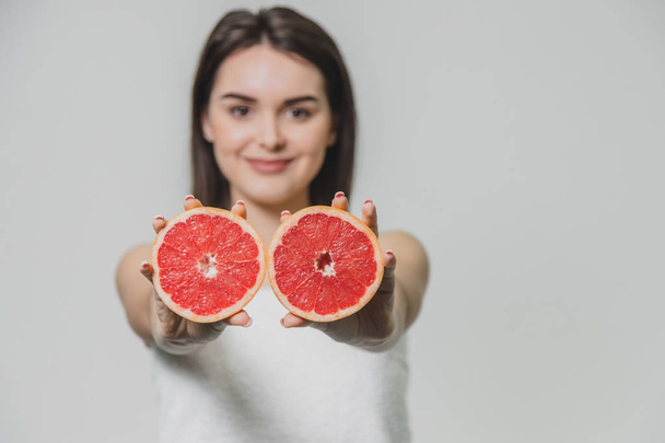 Young sexy girl posing with slices of red grapefruit in her arms extended forward. On a gray background. - Foto, Imagem
