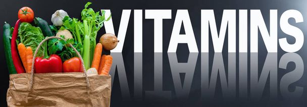 woord vitaminen tegen een donkere achtergrond met verse gezonde biologische groenten in papieren zak - Foto, afbeelding