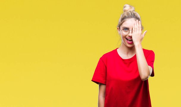 Jovem mulher loira bonita vestindo camiseta vermelha e óculos sobre fundo isolado cobrindo um olho com a mão com sorriso confiante no rosto e emoção surpresa
. - Foto, Imagem