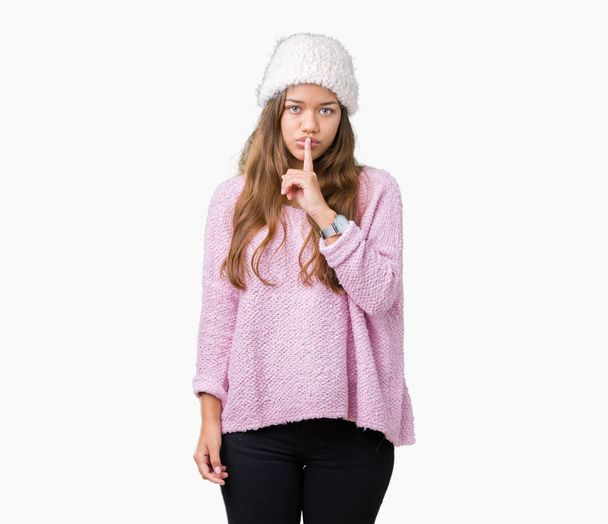 若い美しいブルネット女性の唇に指で静かにするように求めて孤立の背景にセーターと冬の帽子を着ています。沈黙と秘密の概念. - 写真・画像