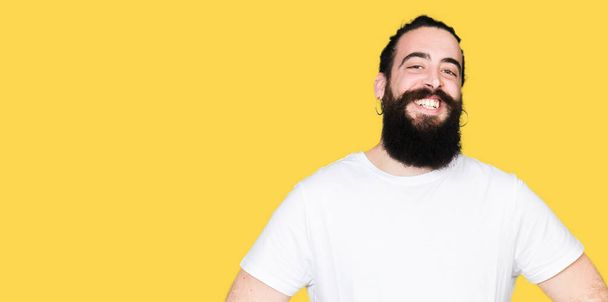 Young hipster man with long hair and beard wearing casual white t-shirt looking away to side with smile on face, natural expression. Laughing confident. - Photo, Image
