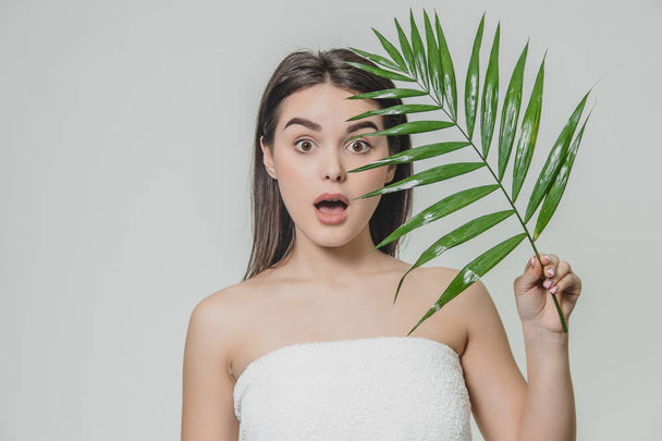 A beautiful young girl in the studio on a white background with wet skin and wet hair holds a large green tropical leaf in hands and covers a part of her face. - Foto, imagen