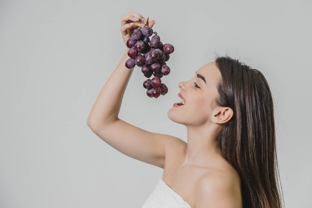 Beautiful young brunette girl with a happy smile. Carrying fresh grapes in hand, isolated on a white background.View from the side. - Foto, Imagem