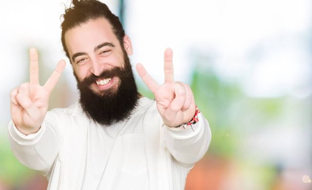 Young man with long hair and beard wearing sporty sweatshirt smiling looking to the camera showing fingers doing victory sign. Number two. - Foto, Imagem