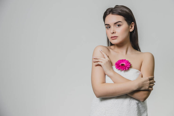 A beautiful young girl with a smooth skin is a light background of natural makeup and curly dark hair. Folding hands around the chest at the level of the chest is a bright flower. - Fotografie, Obrázek