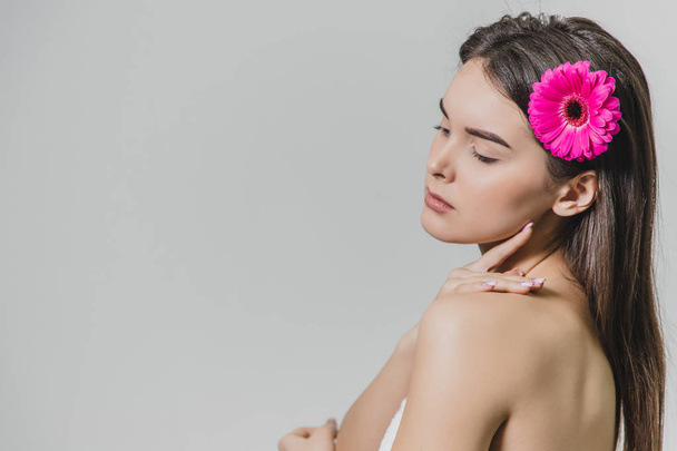 Young pretty girl with a flower in her hair standing on a white background. Has beautiful long black hair. During this, he looks aside. Back view. - Foto, Imagen