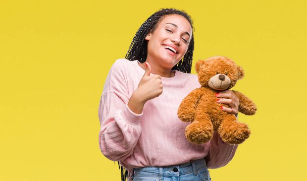 Young african american girl holding teddy bear over isolated background happy with big smile doing ok sign, thumb up with fingers, excellent sign - Foto, Imagem
