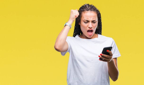 Fiatal fonott hajú afrikai-amerikai lány mutatja a smartphone segítségével elszigetelt háttér, bosszús és csalódott kiabálva a düh, őrült és kiabál, felemelt kézzel, harag koncepció - Fotó, kép