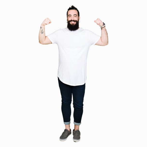 Mladí bokovky muž s dlouhými vlasy a vousy nosí ležérní bílé tričko zobrazeno paže svaly úsměvem hrdá. Fitness koncept. - Fotografie, Obrázek