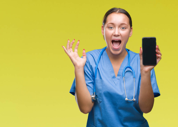 Young caucasian doctor woman showing smarpthone over isolated background very happy and excited, winner expression celebrating victory screaming with big smile and raised hands - Valokuva, kuva