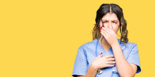 Young adult doctor woman wearing medical uniform smelling something stinky and disgusting, intolerable smell, holding breath with fingers on nose. Bad smells concept. - Fotó, kép
