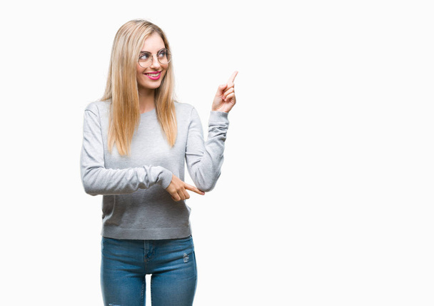 Jonge mooie blonde vrouw bril over geïsoleerde achtergrond met een grote glimlach op het gezicht, met de hand en de vinger te wijzen naar de kant te kijken naar de camera. - Foto, afbeelding