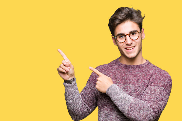 Mladý pohledný muž nosí brýle izolované pozadí s úsměvem a při pohledu na fotoaparát s oběma rukama a prsty přejdete na stranu. - Fotografie, Obrázek