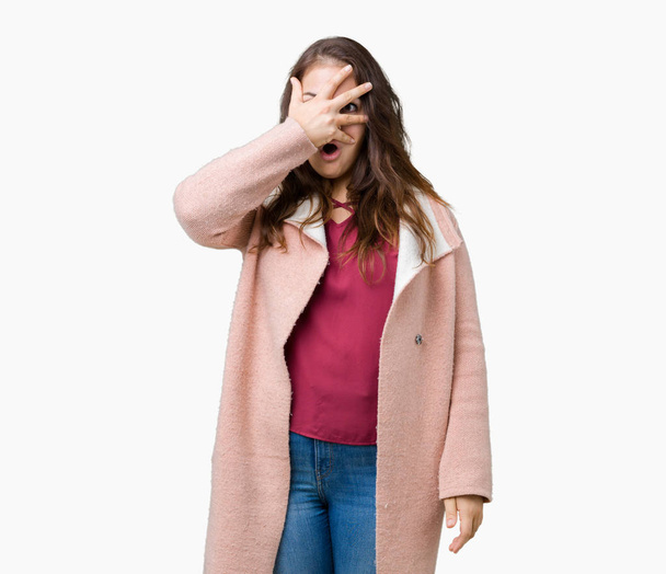 Όμορφη συν μέγεθος νεαρή γυναίκα που φοράει παλτό χειμώνα πέρα από το απομονωμένο υπόβαθρο peeking σε κατάσταση σοκ που καλύπτουν το πρόσωπο και τα μάτια με το χέρι, κοιτάζοντας μέσα από τα δάχτυλα με αμηχανία έκφρασης. - Φωτογραφία, εικόνα