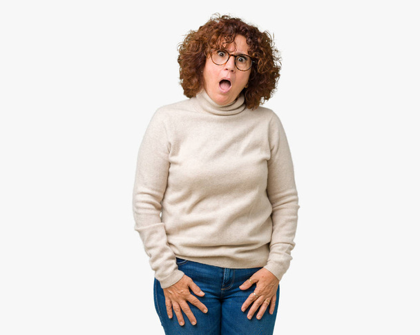 Beautiful middle ager senior woman wearing turtleneck sweater and glasses over isolated background In shock face, looking skeptical and sarcastic, surprised with open mouth - Photo, Image