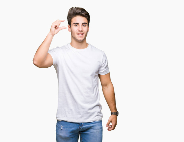 Jonge knappe man dragen witte t-shirt over geïsoleerde achtergrond glimlachend en vertrouwen gebaren met hand grootte teken met vingers doen tijdens het kijken en de camera. Maatregel concept. - Foto, afbeelding