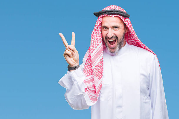 Ανώτερος άνθρωπος Αραβικά φορώντας keffiyeh πέρα από το απομονωμένο υπόβαθρο χαμογελώντας με χαρούμενο πρόσωπο που κλείνει το μάτι στην κάμερα κάνει το σήμα της νίκης. Αριθμός δύο. - Φωτογραφία, εικόνα