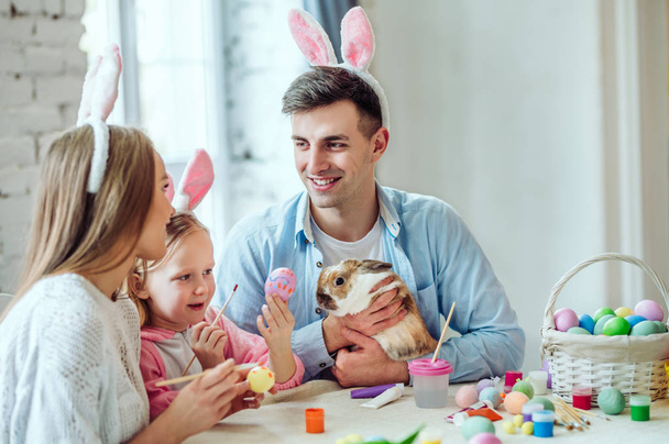 Ми любимо прекрасні традиції. Щаслива мама і дочка фарбують яйця, тато гладить домашнього декоративного кролика
. - Фото, зображення