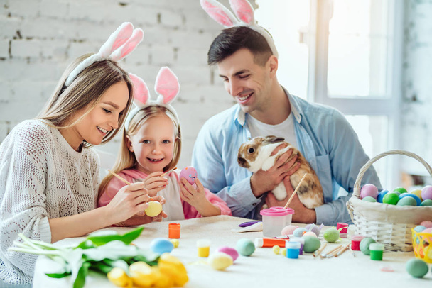 We love beautiful traditions.Mom and daughter paint eggs, dad holds a home decorative rabbit. - Foto, afbeelding