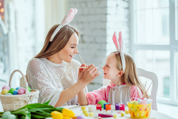 Happy Easter Preparations.Mom and daughter are preparing for Easter together.On the table is a basket with Easter eggs, flowers and paint. - Фото, изображение
