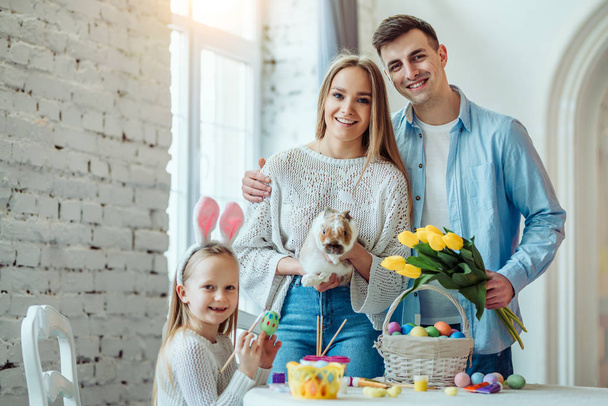 Happy easter!Dad holds a bouquet of tulips, mom holds a home decorative rabbit, daughter paints eggs and looking at camera.Celebration concept. - Фото, изображение