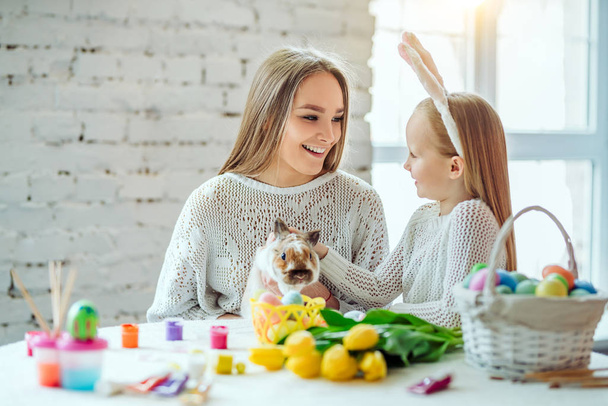 Preparing for Easter with my mother.The little daughter with her mother stroke a home decorative rabbit. - Photo, Image