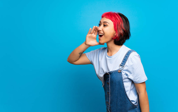 Young woman with pink hair over blue wall shouting with mouth wide open to the lateral - Fotó, kép