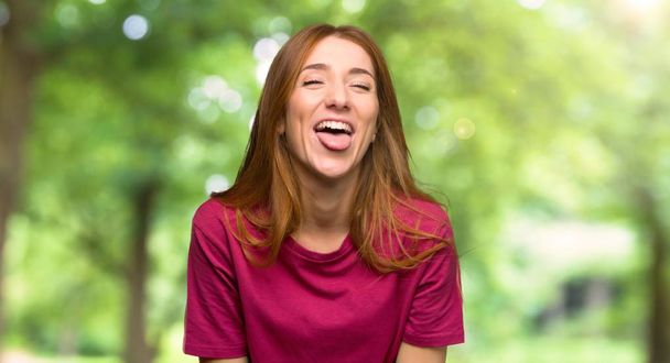 Menina ruiva jovem mostrando língua na câmera com olhar engraçado no parque
 - Foto, Imagem