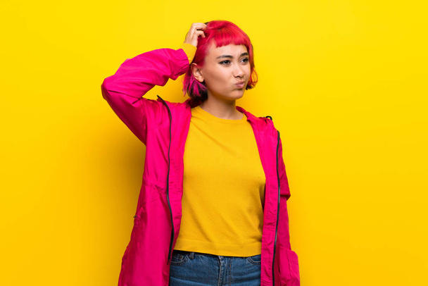 Jeune femme avec les cheveux roses sur le mur jaune ayant des doutes tout en se grattant la tête
 - Photo, image