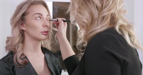 Blonde girl at a beauty salon gets makeup by a professional make up artist - Кадры, видео