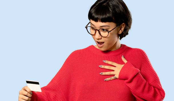 Short hair girl with red sweater holding a credit card and surprised on isolated blue background - Photo, Image