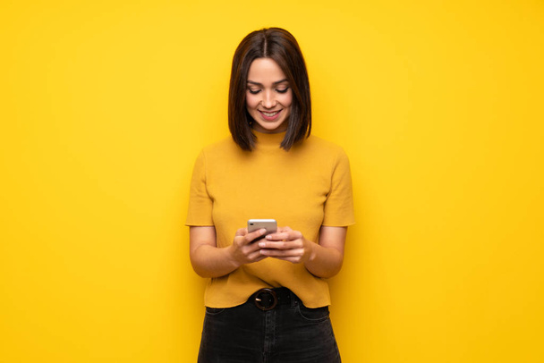 Jonge vrouw over gele muur verzenden van een bericht met de mobiele - Foto, afbeelding