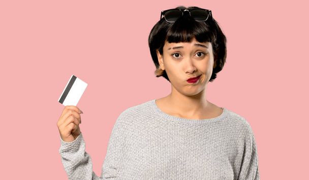 Jonge vrouw met kort haar met een credit card zonder geld op geïsoleerde roze achtergrond - Foto, afbeelding