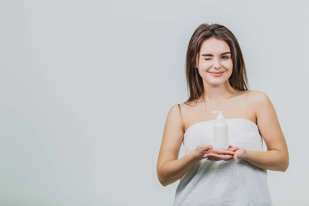 Model look is a beautiful young twin in a white terry towel. Applying a body lotion on her shoulder and smiling while standing on a white background. - Fotoğraf, Görsel