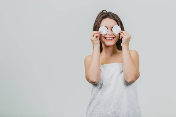 Humorous and positive young beautiful woman with open shoulders covering her eyes with a cotton disc and looking at a smile on a white background. Portrait of happiness. - Foto, Imagem