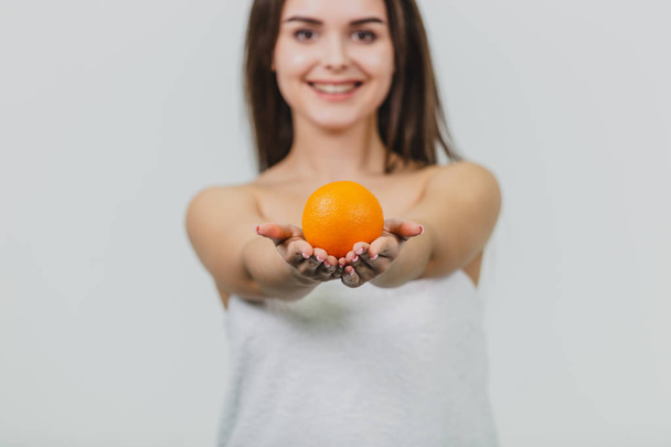 Portrait of a cute young attractive half-eyed woman holding an orange orange on her hands and looking at the camera, isolated over a white background. Orange on the blurred background of the girl. - Photo, image