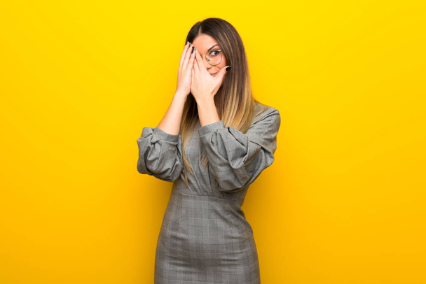 Mujer joven con gafas sobre pared amarilla cubriendo los ojos con las manos y mirando a través de los dedos
 - Foto, Imagen
