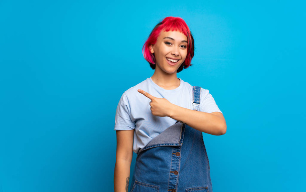 Молода жінка з рожевим волоссям над синьою стіною вказує на бік, щоб представити продукт
 - Фото, зображення