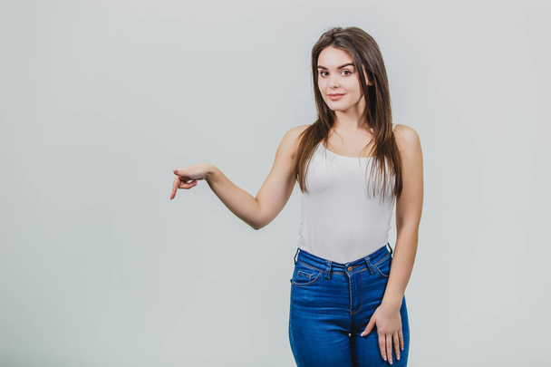 A young pretty Caucasian girl stood on a white background. During this time wear a white T-shirt and blue jeans. Shows the index finger on the floor and opens the mouth to the camera. - Фото, изображение
