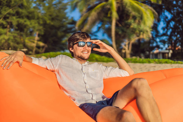 Summer lifestyle portrait of man sitting on the orange inflatable sofa on the beach of tropical island. Relaxing and enjoying life on air bed - Foto, immagini