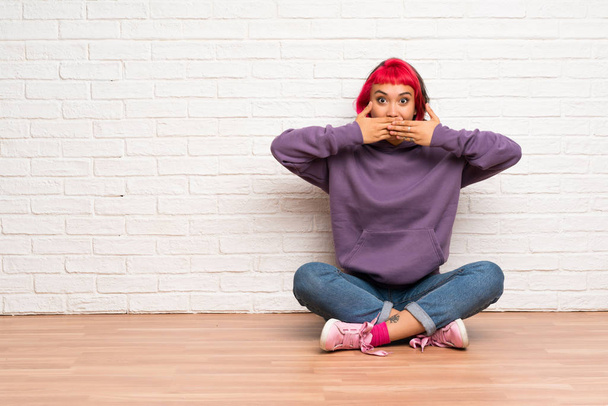 Mladá žena s růžovými vlasy sedí na podlaze pokrývající ústa rukama - Fotografie, Obrázek