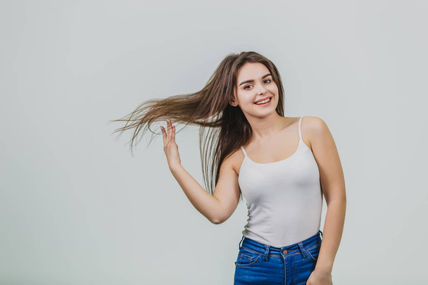 Young pretty girl standing on a white background. It is dressed in white shirt and blue jeans. Has a beautiful long black hair during this throwing it up. - Φωτογραφία, εικόνα