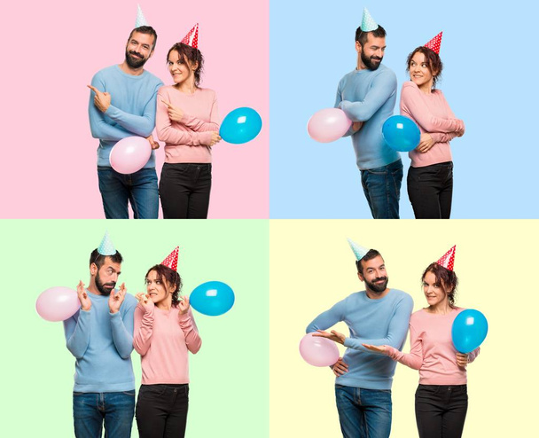 Σύνολο του ζεύγους με τα μπαλόνια και τα καπέλα γενεθλίων που αναζητούν πάνω από τον ώμο με ένα χαμόγελο, επισημαίνοντας την πλευρική και διασχίζουν τα δάχτυλά - Φωτογραφία, εικόνα
