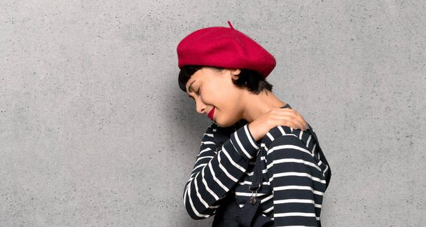 Young woman with beret suffering from pain in shoulder for having made an effort over textured wall - Photo, Image