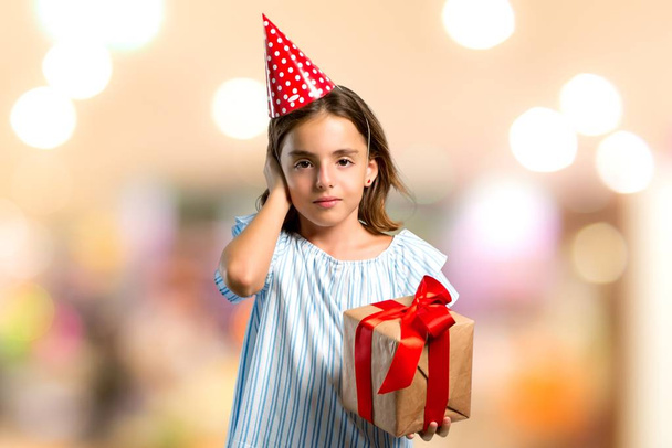 Petite fille à une fête d'anniversaire tenant un cadeau couvrant les deux oreilles avec les mains sur fond flou
 - Photo, image