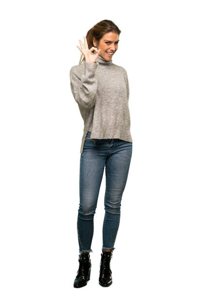 A full-length shot of a Blonde woman with turtleneck showing ok sign with fingers over isolated white background - 写真・画像